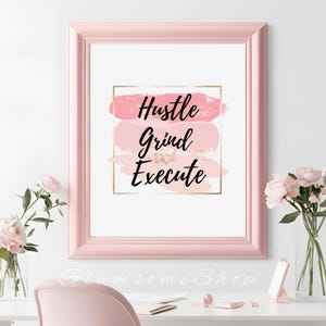 May include: A pink framed print with the words "Hustle Grind Execute" in black text on a white background. The words are layered over a pink and gold abstract design.