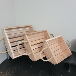 Peut inclure: Trois caisses en bois de tailles différentes, disposées en rangée. Chaque caisse est faite de bois clair avec un design à lattes et des poignées en corde. Les caisses sont vides et semblent neuves.