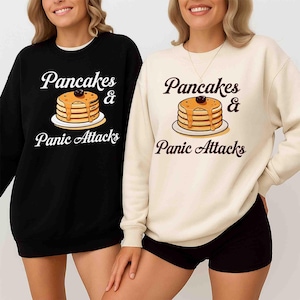 Könnte beinhalten: Zwei Sweatshirts, eines in Schwarz und eines in Creme, jeweils mit dem Text "Pancakes & Panic Attacks" über einer Illustration von Pfannkuchen mit Sirup und einer Kirsche. Das schwarze Sweatshirt hat einen weißen Kragen.