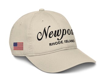 May include: A beige baseball cap with an embroidered American flag on the left side and the text "Newport Rhode Island" in black cursive script.