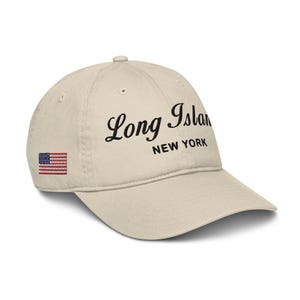May include: A beige baseball cap with a black embroidered design that says "Long Island New York" and a small American flag patch on the side.