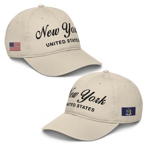 May include: Two beige baseball caps with curved visors. The caps feature the words "New York UNITED STATES" in black script. One cap has a small embroidered American flag, the other has a New York State flag.