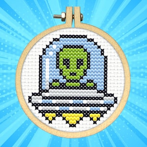 Puede incluir: Bordado de punto de cruz de un extraterrestre verde en un OVNI, en un aro de madera. El extraterrestre tiene grandes ojos negros y está rodeado por un escudo azul claro. El OVNI es blanco y negro con detalles amarillos.