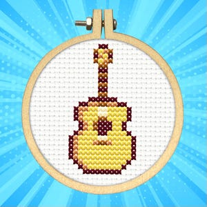 May include: A cross-stitch embroidery of a yellow and brown guitar within a wooden embroidery hoop. The guitar design features a detailed body and neck, with dark brown accents. The background is a white fabric with a blue radial burst.