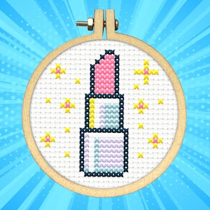 May include: A cross-stitch embroidery of a lipstick in a wooden hoop. The lipstick is pink and the base is a mix of blue, yellow, and purple. Yellow starbursts surround the lipstick on a white background.