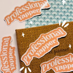Könnte beinhalten: Vier pfirsichfarbene Aufkleber mit dem Text "Professional Yapper" in weißen Buchstaben. Die Aufkleber befinden sich auf einem braunen Korkbrett und einer blau-weißen gemusterten Oberfläche.
