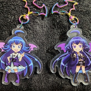 May include: Two clear acrylic charms featuring anime-style characters with purple and blue hair. The characters are wearing outfits with a gothic theme. The charms are attached to a silver chain with a bat-shaped design.