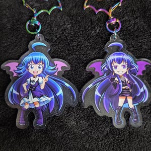 May include: Two acrylic keychains depicting anime-style characters with blue hair and purple bat wings. The keychains have iridescent bat-shaped top attachments and silver clasps. One character wears a purple dress, the other a black outfit.