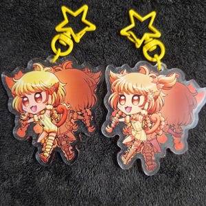May include: Two clear acrylic keychains featuring a cartoon character with yellow hair and a yellow outfit. The character has cat-like features and is smiling. Each keychain has a yellow star-shaped clasp.