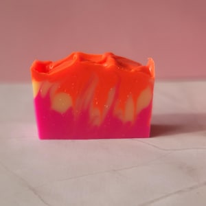 May include: A vibrant bar of handmade soap with a layered design. The bottom layer is bright pink, transitioning to yellow and then a swirled orange top. The soap is rectangular and sits on a white surface with a pink background.
