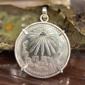 May include: Silver pendant featuring a 1933 Mexican peso coin with the inscription "UN PESO 1933" and a sunburst design on the front.