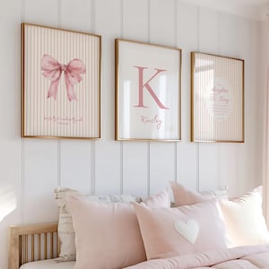 May include: Three framed wall art pieces with gold frames. The art features a pink bow, a pink letter "K" with the name "Kinsley", and a circular design with text. The art hangs above a bed with pink bedding and pillows.