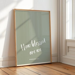 May include: A framed print with a sage green background and white text that reads "How blessed are we" with the scripture reference "Ephesians 1:3-4" below.
