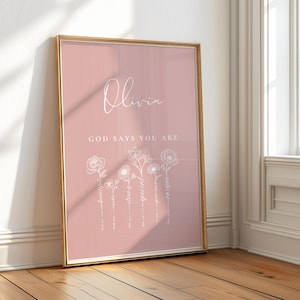 Puede incluir: Una impresión enmarcada rosa con el nombre "Olivia" y el texto "Dios dice que eres" con ilustraciones florales blancas.