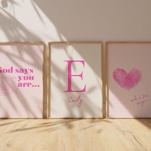 May include: Three framed art prints with pink and cream backgrounds. The left print has the text "God says you are...". The centre print features a pink letter "E" and the name "Emily". The right print shows a pink fingerprint heart.