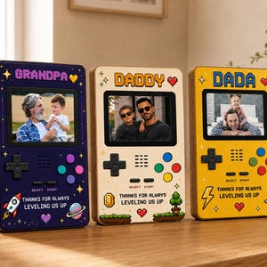 May include: Three wooden photo frames shaped like retro handheld game consoles. The frames are navy blue, beige, and yellow, each with a different family member's name at the top. Each frame has a photo inside and the text "THANKS FOR ALWAYS LEVELING US UP".