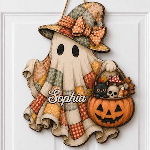 May include: A hanging wall decoration shaped like a ghost wearing a patchwork hat with a bow. The ghost features the name Sophia, and holds a pumpkin basket containing a black cat, a skull, and mushrooms. The design uses orange, tan, and green.