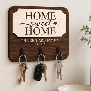 May include: A rectangular dark wood wall-mounted key holder with a light wood plaque featuring the text "HOME sweet HOME" and a heart icon. Below, the text reads "THE RICHARD FAMILY" and "EST. 2026". Three black metal hooks hold various keys.