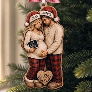 Puede incluir: Un adorno navideño de madera con una pareja en pantalones de pijama de cuadros rojos y suéteres color crema. La persona de la izquierda sostiene una ecografía. Ambos llevan gorros de Papá Noel con "Jean" y "James". Un corazón grabado "2026".