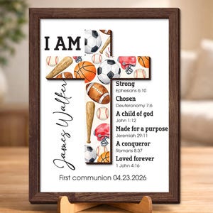 May include: A framed print with a cross design, incorporating sports equipment like baseballs, footballs, and soccer balls. The print includes the text "I AM" and inspirational phrases. The frame is brown, and the print is displayed on a wooden stand.