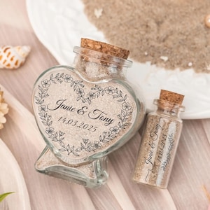 Pode incluir: Duas garrafas de vidro cheias de areia, uma em forma de coração e outra pequena, ambas com rolhas de cortiça. A garrafa em forma de coração tem os nomes "Janie & Tony" e a data "14.03.2025" dentro de uma grinalda floral. Lembrancinhas de casamento.