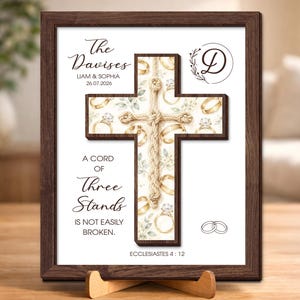 May include: A framed wall art piece featuring a central wooden cross with a rope overlay. The background displays gold wedding rings and greenery. Text includes The Davises, Liam & Sophia, 26.07.2026, A Cord of Three Strands Is Not Easily Broken, and Ecclesiastes 4:12.