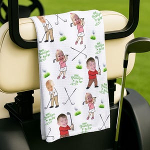 May include: White golf towel with a repeating pattern of children playing golf, golf clubs, golf balls, and the text "Best Grandpa By Par" in green. The towel is hanging on a golf cart.