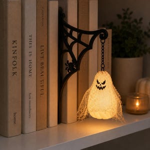 May include: A black metal wall bracket featuring a spider web design with a small spider, holding a hanging light fixture shaped like a ghost. The ghost is covered in a thin, off-white mesh fabric and displays a dark, jagged facial expression.