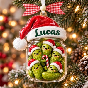 Könnte beinhalten: Ein durchsichtiger Glasanhänger in Form eines Einmachglases mit dem Namen Lucas in grüner Schrift auf dem weißen Rand einer Weihnachtsmütze. Im Glas befinden sich mehrere grüne, lächelnde Gewürzgurken, die jeweils eine kleine rot-weiße Weihnachtsmütze tragen. Eine Gurke hält eine rot-weiße Zuckerstange, und oben ist eine rot-weiß karierte Schleife befestigt.