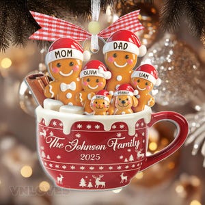 Könnte beinhalten: Ein Weihnachtsschmuck in Form einer roten Tasse, gefüllt mit Lebkuchenfiguren und Haustieren mit Weihnachtsmützen. Die Tasse zeigt weiße Rentier- und Schneeflockenmuster, den Text The Johnson Family 2025 und eine rot-weiße Vichy-Schleife.