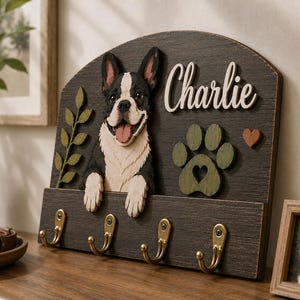 K&ouml;nnte beinhalten: Ein Schl&uuml;sselhalter aus dunklem Holz zur Wandmontage mit einem 3D-Ausschnitt einer schwarz-wei&szlig;en Franz&ouml;sischen Bulldogge, einem gr&uuml;nen Pfotenabdruck, einem kleinen braunen Herz und dem Namen Charlie in wei&szlig;er Schrift sowie vier Messinghaken.