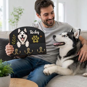 K&ouml;nnte beinhalten: Ein Wand-Schl&uuml;sselhalter aus dunklem Holz mit einem gemalten Siberian Husky, einem goldenen Pfotenabdruck, einem kleinen Herz und dem Namen Rocky in wei&szlig;er Schrift. Die Platte hat drei Haken aus messingfarbenem Metall.
