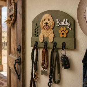 K&ouml;nnte beinhalten: Ein wandmontiertes, olivgr&uuml;nes Holzhakenbrett mit einem skulptierten goldbraunen Hund, einer gelben Pfote und dem Namen Buddy in wei&szlig;er Schrift. Vier schwarze Metallhaken halten eine Leine, ein Lederhalsband, einen Riemen und Schl&uuml;ssel.