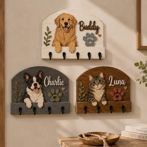 K&ouml;nnte beinhalten: Drei wandmontierte Schl&uuml;sselhalter aus Holz mit geschnitzten Tierfiguren. Ein wei&szlig;es Brett zeigt einen Golden Retriever namens Buddy, ein graues Brett eine Franz&ouml;sische Bulldogge namens Charlie und ein braunes Brett eine getigerte Katze namens Luna.