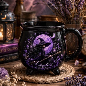 May include: A black ceramic cauldron-shaped mug featuring a graphic of a witch in a pointed hat holding a broomstick against a large purple moon. The design includes purple floral accents and the name Elizabeth printed in a white script font.