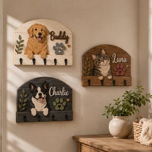 K&ouml;nnte beinhalten: Drei bogenf&ouml;rmige Wandorganizer aus Holz mit geschnitzten Tierfiguren. Sie zeigen einen Golden Retriever namens Buddy, einen Boston Terrier namens Charlie und eine getigerte Katze namens Luna, jeweils mit vier schwarzen Haken und Pfotenabdr&uuml;cken.