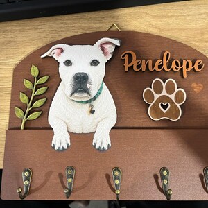 K&ouml;nnte beinhalten: Ein h&ouml;lzernes Wand-Schl&uuml;sselbrett mit einem wei&szlig;en Hund mit gr&uuml;nem Halsband, einem gr&uuml;nen Blattzweig, einem Pfotenabdruck-Ausschnitt und dem Namen Penelope in Schreibschrift. Das Brett hat f&uuml;nf messingfarbene Metallhaken an der Unterkante.