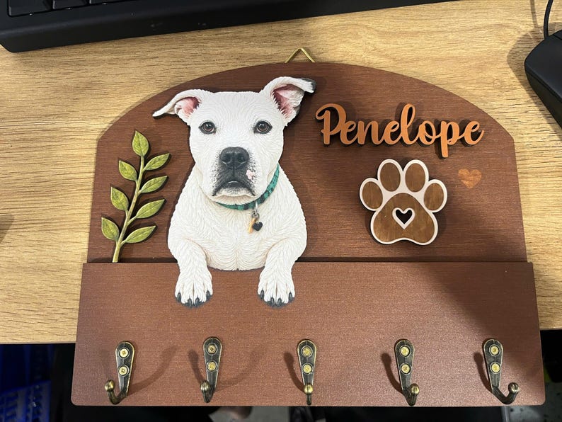 K&ouml;nnte beinhalten: Ein h&ouml;lzernes Wand-Schl&uuml;sselbrett mit einem wei&szlig;en Hund mit gr&uuml;nem Halsband, einem gr&uuml;nen Blattzweig, einem Pfotenabdruck-Ausschnitt und dem Namen Penelope in Schreibschrift. Das Brett hat f&uuml;nf messingfarbene Metallhaken an der Unterkante.