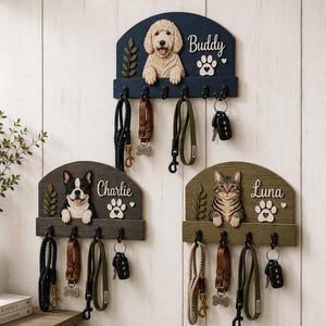 K&ouml;nnte beinhalten: Drei wandmontierte Leinen- und Schl&uuml;sselhalter aus Holz mit geschnitzten Figuren eines Golden Doodles, einer Franz&ouml;sischen Bulldogge und einer getigerten Katze. Jedes Brett zeigt einen Namen, eine Pfote und einen Zweig mit f&uuml;nf schwarzen Haken.