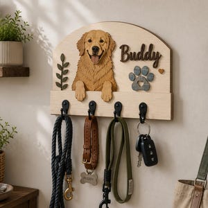 K&ouml;nnte beinhalten: Ein helles Wand-Schl&uuml;sselbrett aus Holz mit einem dreidimensionalen Ausschnitt eines Golden Retrievers, einem kleinen gr&uuml;nen Blattzweig, einem dunkelbraunen Namensschild mit der Aufschrift Buddy und einer grauen Pfote mit Herz.