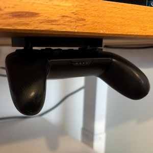 Könnte beinhalten: Ein schwarzer Videospiel-Controller ist mit einer schwarzen Plastikhalterung unter einem Holztisch montiert. Der Controller hängt kopfüber, die Tasten zeigen zur Decke.