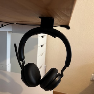 Könnte beinhalten: Schwarzer Over-Ear-Kopfhörer, der an einem schwarzen Kopfhörerhalter unter dem Schreibtisch hängt. Der Kopfhörer hat ein eingebautes Mikrofon und hängt unter einem Holzschreibtisch. Der Hintergrund umfasst einen weißen Schrank und eine beige Wand.