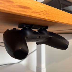 Könnte beinhalten: Ein schwarzer Videospielcontroller ist unter einem Holztisch montiert. Der Controller hängt kopfüber mit den Knöpfen nach oben.