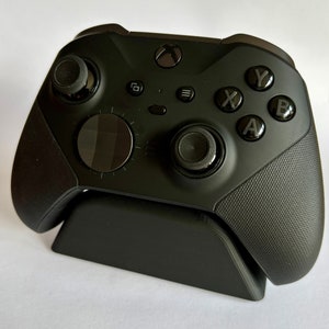 Könnte beinhalten: Ein schwarzer Xbox-Controller mit einem Ständer. Der Controller hat eine strukturierte Oberfläche und verfügt über Tasten mit den Beschriftungen A, B, X und Y. Der Controller steht auf einem schwarzen Ständer.