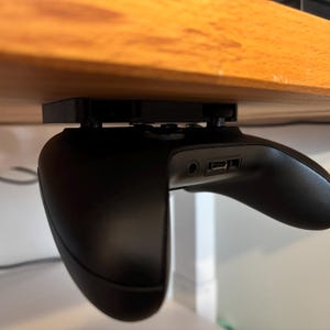 Könnte beinhalten: Ein schwarzer Videospielcontroller ist mit einer 3D-gedruckten Halterung unter einem Holztisch montiert. Der Controller hängt kopfüber mit den Knöpfen zum Tisch.