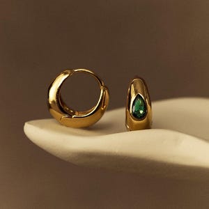 Può includere: Un paio di orecchini a cerchio color oro. Un orecchino presenta una gemma verde smeraldo a forma di goccia. Gli orecchini sono esposti su una superficie bianca su uno sfondo caldo e neutro. Il logo The Knot KnotCo è visibile.