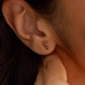 Może przedstawiać: Zbliżenie ucha z kolczykiem w kształcie złotej laski cukrowej. Kolczyk jest ozdobiony czarnymi i czerwonymi kamieniami. W prawym dolnym rogu widnieje napis The Knot Knot Co.