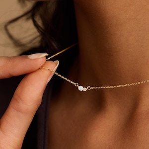 Puede incluir: Un delicado collar de oro con una pequeña ópalo y un diminuto diamante. El collar se sostiene, mostrando su fina cadena y diseño minimalista. The Knot Knot Co. está impreso en la parte inferior derecha.