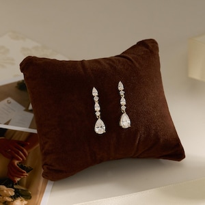May include: A pair of gold-toned drop earrings featuring three clear, faceted, pear-shaped and round-cut stones arranged vertically. The earrings are displayed on a dark brown, square-shaped velvet cushion with a soft, textured surface.