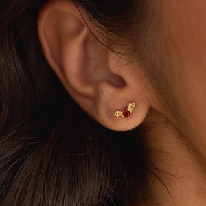 May include: Gold-colored stud earrings with a bat wing design. Each earring features a heart-shaped, red gemstone at the center. The earrings are worn on a person's ear. The Knot Knot Co. is written in the bottom right corner.